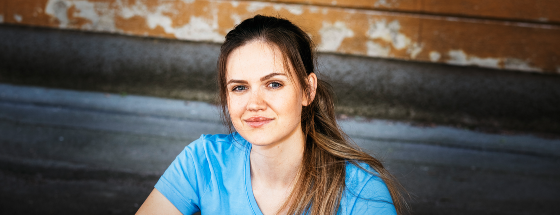 Caritas Pflege: Wir sind die Antwort auf Who Cares. Eine junge Frau mit braunen langen Haaren und Pferdeschwanz sitzt auf einer Stiege, sie trägt ein hellblaues T-Shirt mit Caritas Logo und lächelt in die Kamera.